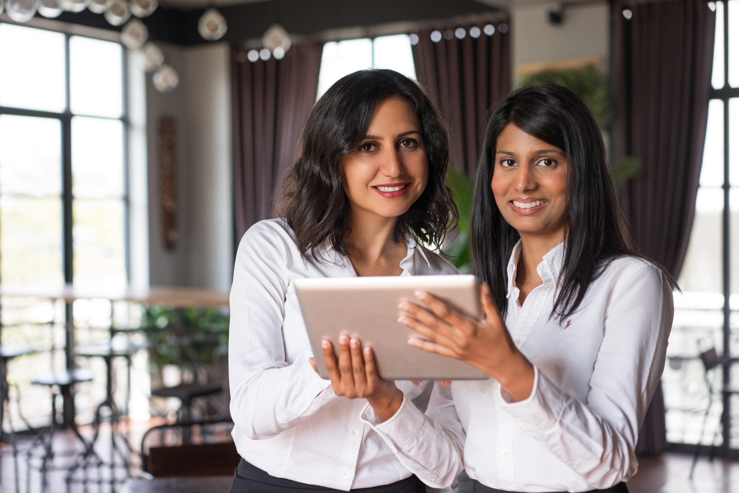 Two smiling female coworkers using tablet computer in cafe. They are standing and looking at camera with cafe interior and windows in background. Technology concept. Front view.